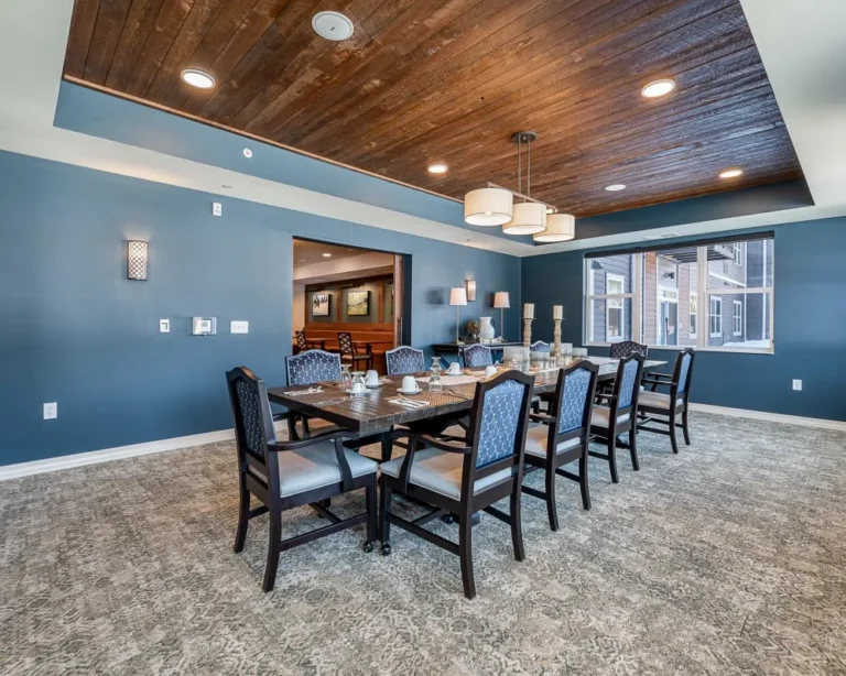 A long wooden dining table set for a meal in a blue-walled room with a dark wood ceiling and circular pendant lights.