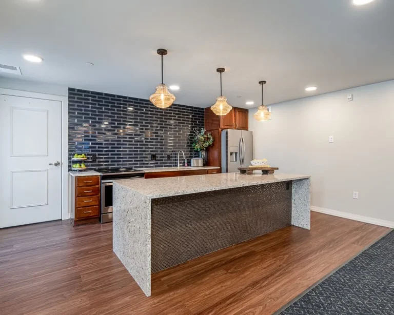 Modern kitchen with a large speckled island, dark subway tile backsplash, pendant lights, wooden cabinets, and stainless steel fridge.