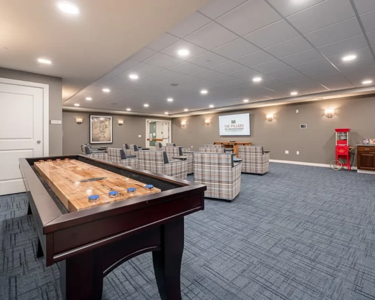 Recreation room with a wooden shuffleboard table in the foreground and plaid armchairs arranged around a central seating area.